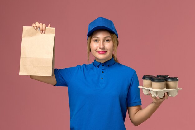 Courrier féminin en uniforme bleu tenant un paquet de nourriture et des tasses de livraison marron sur rose, livreur d'uniforme de service