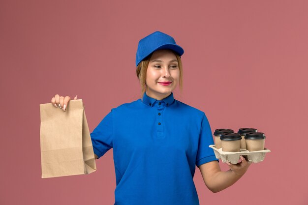 Courrier féminin en uniforme bleu tenant un paquet de nourriture et des tasses de livraison marron sur rose, livreur d'uniforme de service