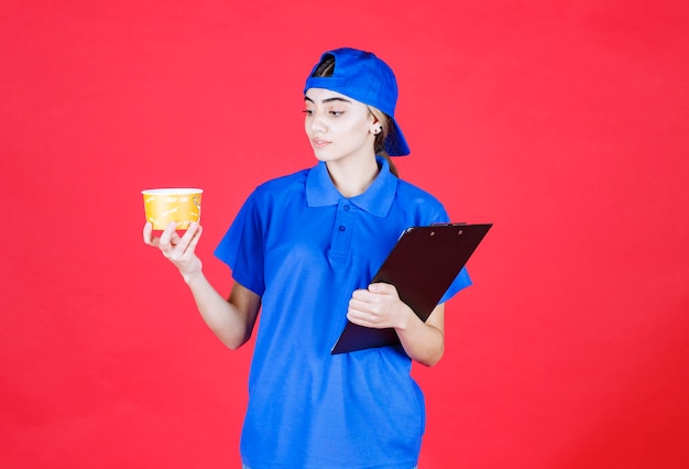 Courrier féminin en uniforme bleu tenant une tasse de nouilles jaunes et un dossier noir.