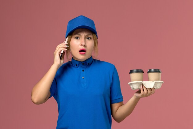 Courrier féminin en uniforme bleu tenant des tasses de café de livraison et parler au téléphone sur rose, livraison uniforme de travail de travailleur de service