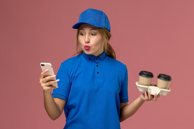 Photo gratuite courrier féminin en uniforme bleu tenant des tasses de café de livraison et en utilisant son téléphone sur rose, la livraison d'uniforme de travailleur de service