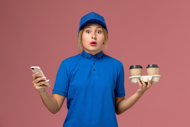 Courrier féminin en uniforme bleu tenant des tasses de café de livraison et en utilisant son téléphone sur rose, la livraison d'uniforme de travailleur de service