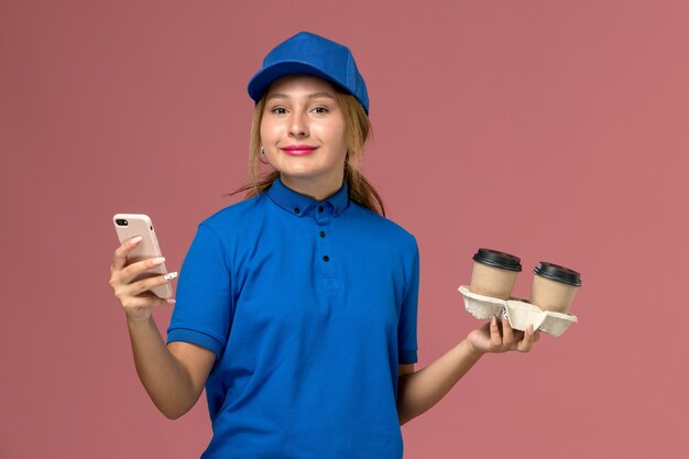 Courrier féminin en uniforme bleu tenant des tasses de café de livraison et en utilisant son téléphone avec le sourire sur rose, la livraison d'uniforme de travailleur de service