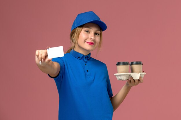 Courrier féminin en uniforme bleu tenant des tasses de livraison de café et carte blanche avec sourire sur rose, livreur d'uniforme de service