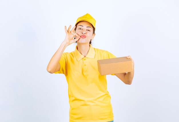 Courrier féminin en uniforme jaune livrant un colis en carton et montrant un signe de main réussi.