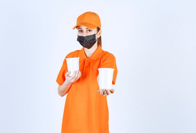 Courrier féminin en uniforme jaune et masque noir tenant des boissons à emporter dans des gobelets en plastique à deux mains et les offrant au client.