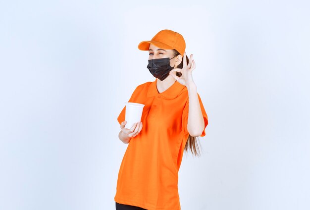 Photo gratuite courrier féminin en uniforme jaune et masque noir tenant une tasse à emporter en plastique et appréciant le goût.