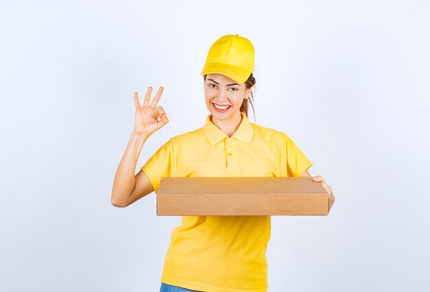 Courrier féminin en uniforme jaune tenant un colis en carton et montrant un signe de la main de plaisir.