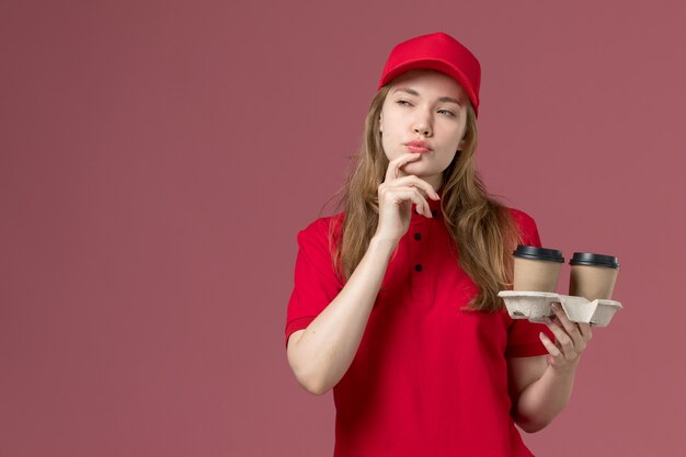 Courrier féminin en uniforme rouge avec expression de la pensée tenant des tasses à café sur rose, travailleur de l'emploi de livraison de service uniforme