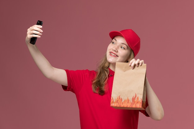 Courrier féminin en uniforme rouge prenant selfie holding paper food package sur rose clair, travail uniforme de livraison des travailleurs des services