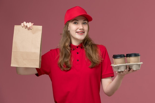 Courrier féminin en uniforme rouge souriant tenant des tasses de café de livraison marron et paquet de nourriture sur rose, travailleur de travail de livraison de service uniforme