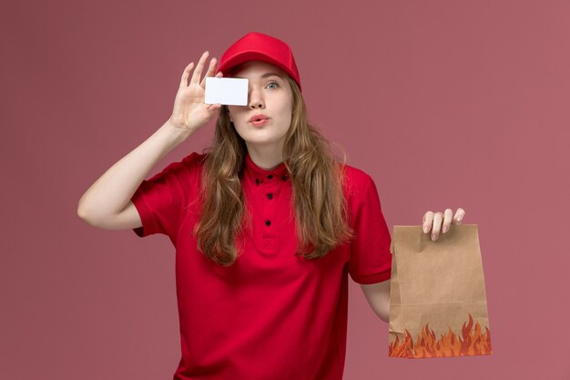 Courrier féminin en uniforme rouge tenant un paquet de papier alimentaire avec carte blanche sur le rose clair, la prestation de services de travail uniforme de travail