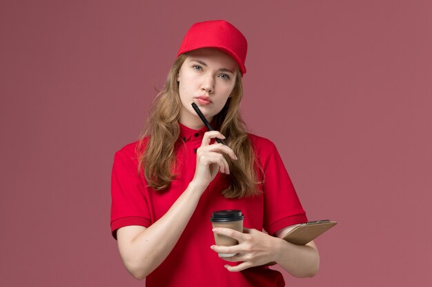 Courrier féminin en uniforme rouge tenant des tasses de café bloc-notes et stylo pensant sur rose, travailleur de travail de livraison de service uniforme