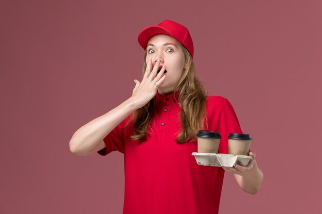 Courrier féminin en uniforme rouge tenant des tasses à café avec expression surprise sur rose, travail de prestation de services uniforme