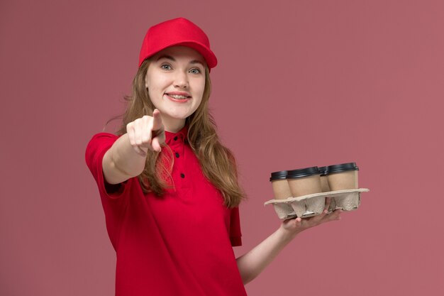 Courrier féminin en uniforme rouge tenant des tasses de café de livraison avec un large sourire sur rose, prestation de services de travailleur uniforme de travail