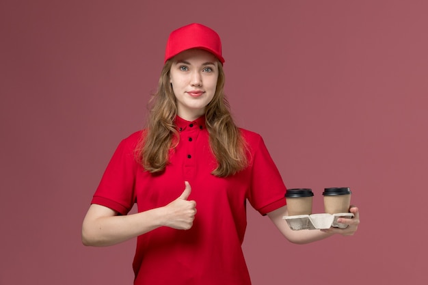 Courrier féminin en uniforme rouge tenant des tasses de café de livraison marron posant sur le rose, travailleur de service d'emploi uniforme