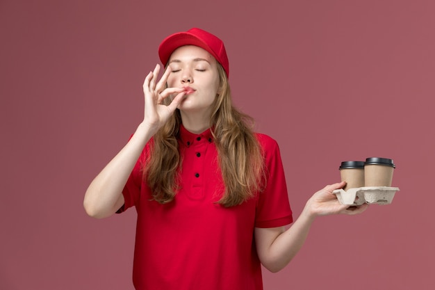 Courrier féminin en uniforme rouge tenant des tasses de café de livraison sur rose clair, travail uniforme de livraison des travailleurs de service