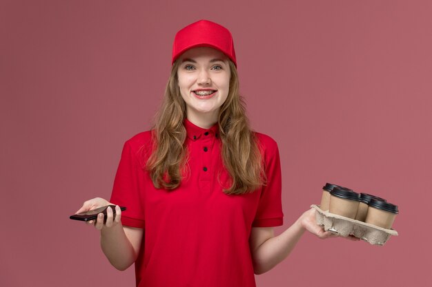 Courrier féminin en uniforme rouge tenant des tasses de café de livraison et téléphone souriant sur rose, prestation de services de travail uniforme