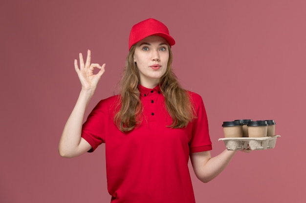 Courrier féminin en uniforme rouge tenant des tasses de café marron sur rose clair, travail uniforme de livraison des travailleurs des services