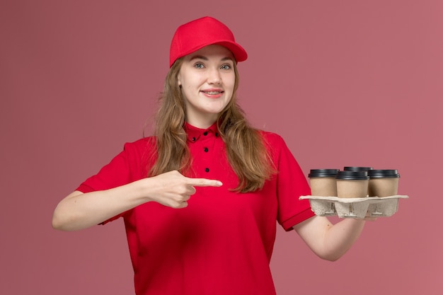 Courrier féminin en uniforme rouge tenant des tasses à café avec sourire sur rose clair, travail uniforme de livraison des travailleurs