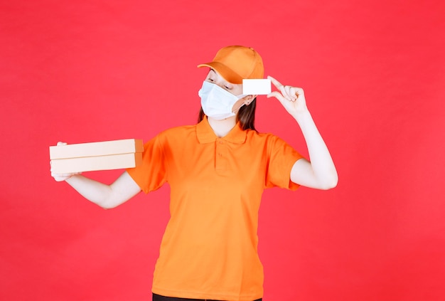 Photo gratuite coursier féminin en code vestimentaire de couleur orange et masque tenant une boîte en carton et présentant sa carte de visite.