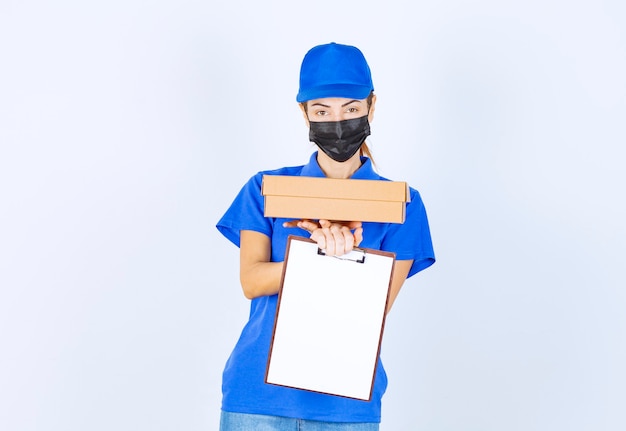 Coursière en uniforme bleu et masque facial livrant un colis en carton et demandant au client de signer sur le blanc.