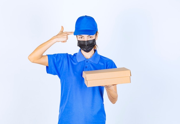 Coursière en uniforme bleu et masque facial tenant une boîte en carton et pensant à la livraison.