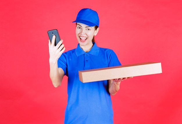 Coursière en uniforme bleu tenant une boîte à pizza à emporter en carton et prenant son selfie.