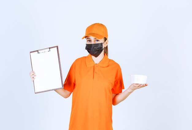 Coursière en uniforme orange et masque noir tenant un gobelet en plastique et une liste de clients