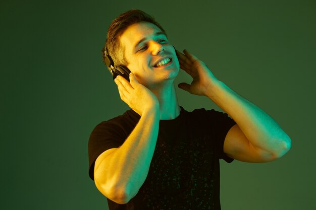 Écouter de la musique, danser, chanter. Portrait de l'homme caucasien sur fond de studio vert en néon. Beau modèle masculin en chemise noire. Concept d'émotions humaines, expression faciale, ventes, publicité.