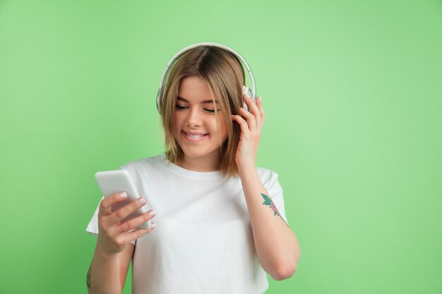 Écouter de la musique. Portrait de jeune femme caucasienne isolé sur mur vert. Beau modèle féminin en chemise blanche.