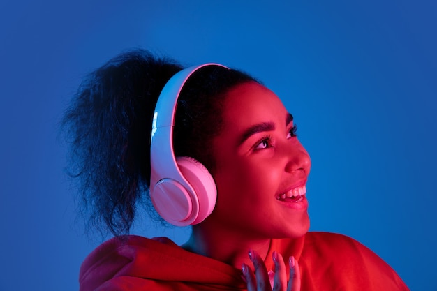 Écouteurs. Portrait de femme afro-américaine isolé sur fond bleu studio en néon multicolore. Beau modèle féminin. Concept d'émotions humaines, expression faciale, ventes, publicité, mode.