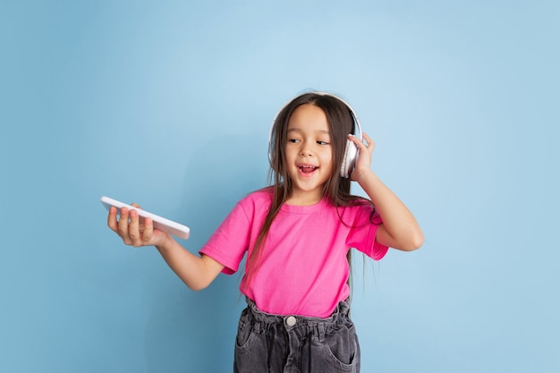 Écoutez le portrait d'une petite fille caucasienne musi sur un mur bleu. Beau modèle féminin en chemise rose. Concept d'émotions humaines, expression faciale, jeunesse, enfance.