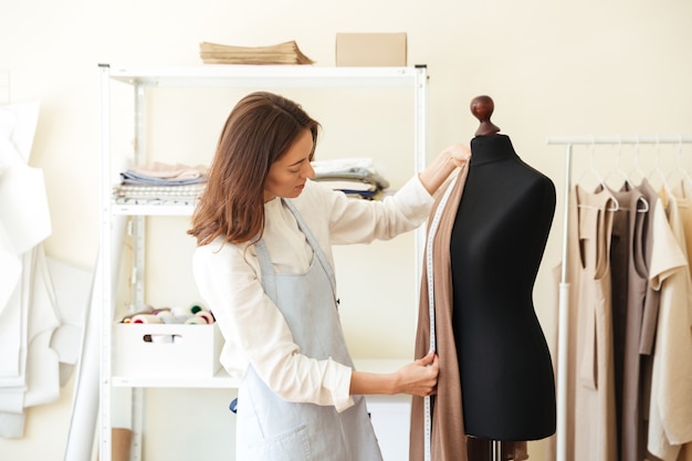 Couturière brune en tablier mesurant un beau tissu sur un mannequin noir en atelier