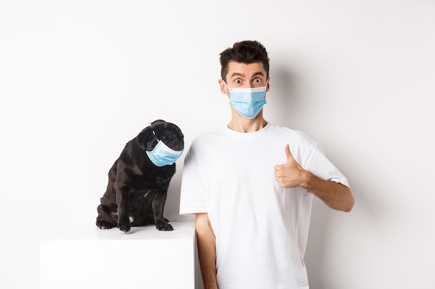 Covid-19, animaux et concept de quarantaine. Image d'un jeune homme drôle et d'un petit chien portant des masques médicaux, le propriétaire montrant le pouce en l'air en signe d'approbation, loue quelque chose, fond blanc