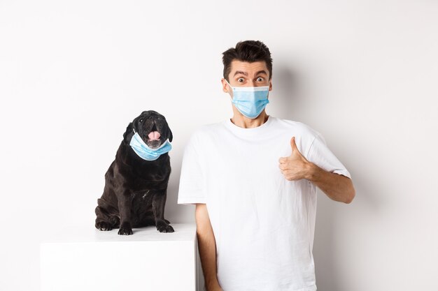 Covid-19, animaux et concept de quarantaine. Image d'un jeune homme drôle et d'un petit chien portant des masques médicaux, le propriétaire montrant le pouce en l'air en signe d'approbation, loue quelque chose, fond blanc