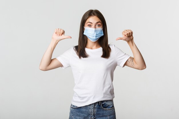 Covid-19, concept de distanciation sanitaire et sociale. Jolie femme souriante en masque médical pointant sur elle-même, blanc.