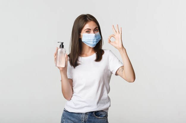 Covid-19, concept de distanciation sanitaire et sociale. Portrait de jeune fille brune souriante dans un masque médical, montrant un désinfectant pour les mains et un geste correct.