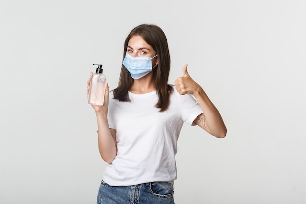 Covid-19, concept de distanciation sanitaire et sociale. Portrait de jeune fille souriante satisfaite en masque médical, montrant le pouce en l'air et désinfectant pour les mains.