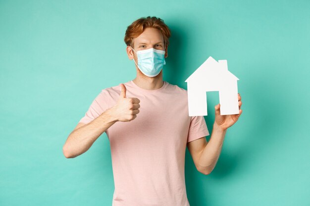 Covid-19 et concept immobilier. Un gars joyeux en masque facial montrant une découpe de maison et un pouce levé, debout sur fond turquoise