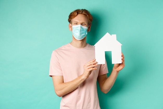 Covid-19 et concept immobilier. Jeune homme heureux en masque facial montrant la découpe de maison en papier et souriant, offre une propriété à vendre, debout sur fond de menthe.