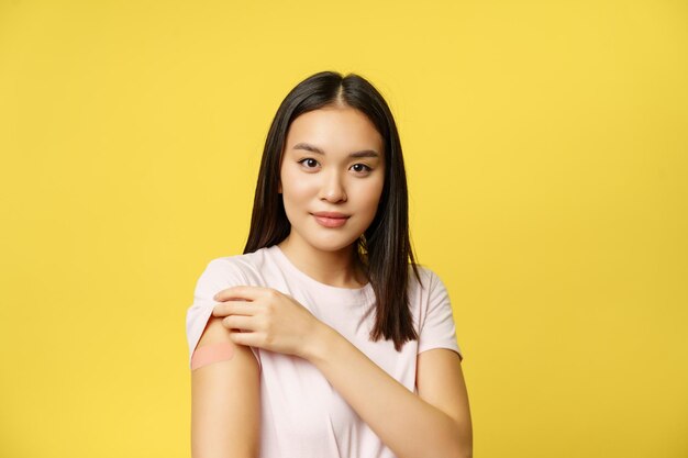 Covid-19 et concept médical de soins de santé. Une fille coréenne souriante montre une épaule avec un patch, a pris une photo du vaccin contre le coronavirus pendant la pandémie, fond jaune.