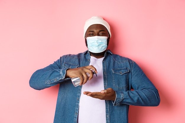 Covid-19, concept de mode de vie et de verrouillage. Homme afro-américain souriant au masque facial nettoyant les mains avec un désinfectant, utilisant un antiseptique et regardant la caméra, fond rose.