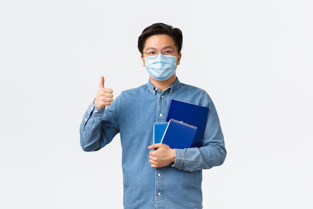 Covid-19, prévention du virus et distanciation sociale au concept universitaire. Un enseignant ou un tuteur asiatique joyeux portant un masque médical porte des cahiers et du matériel d'étude, montrant le pouce levé, fond blanc.