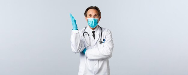 Covid-19, prévention du virus, travailleurs de la santé et concept de vaccination. Médecin de sexe masculin caucasien excité portant un masque médical et des gants, levez la main, ayez une idée, plan de réflexion, fond blanc.