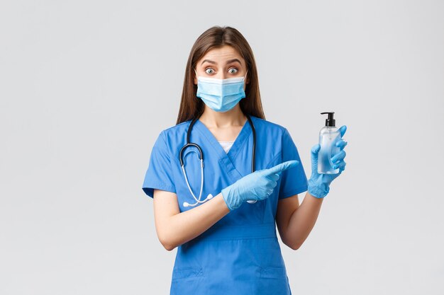 Covid-19, prévention des virus, santé, travailleurs de la santé et concept de quarantaine. Infirmière ou médecin enthousiaste en gommage bleu, masque médical et gants, pointant vers le désinfectant pour les mains, a l'air surpris