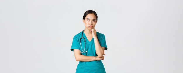Covid-19, travailleurs de la santé, concept de pandémie. Femme médecin asiatique agacée et dérangée, infirmière pendant son quart de travail ayant l'air irritée ou fatiguée, roulement des yeux ennuyé et expiré, debout sur fond blanc dans les gommages