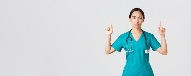 Covid-19, travailleurs de la santé, concept de pandémie. Sombre femme médecin asiatique déçue, médecin en gommage pointant les doigts vers le haut et grimaçant mécontent, debout sur fond blanc