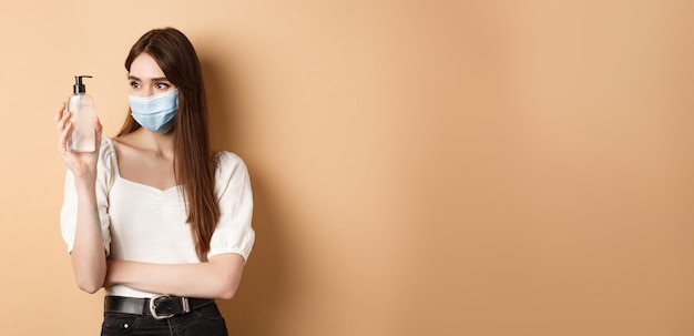 Photo gratuite covid et concept de mesures préventives une fille souriante dans un masque facial regarde une bouteille de désinfectant pour les mains en utilisant