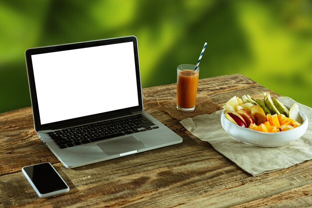 Écrans vierges d'ordinateur portable et de smartphone sur une table en bois à l'extérieur avec la nature sur le mur Fruits et jus de fruits frais à proximité. Concept de lieu de travail créatif, entreprise, indépendant. Copyspace.